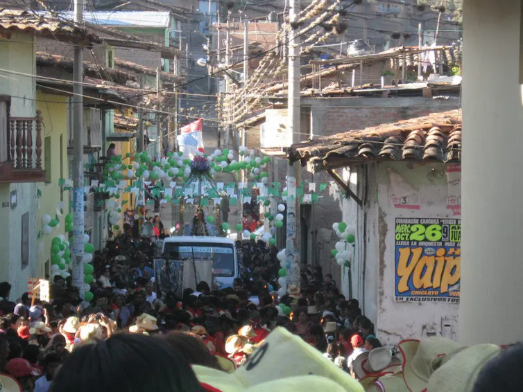 A white van moves through a street that is packed with people and festooned with green and white balloons.