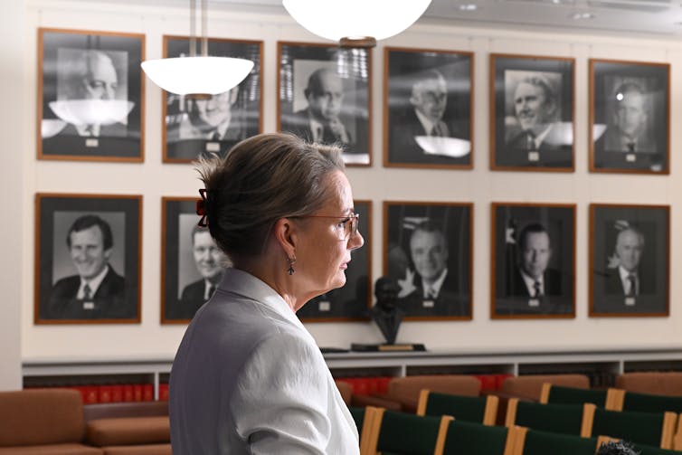 Sussan Ley in a white jacket standing side on before a wall of black and whitephotographs