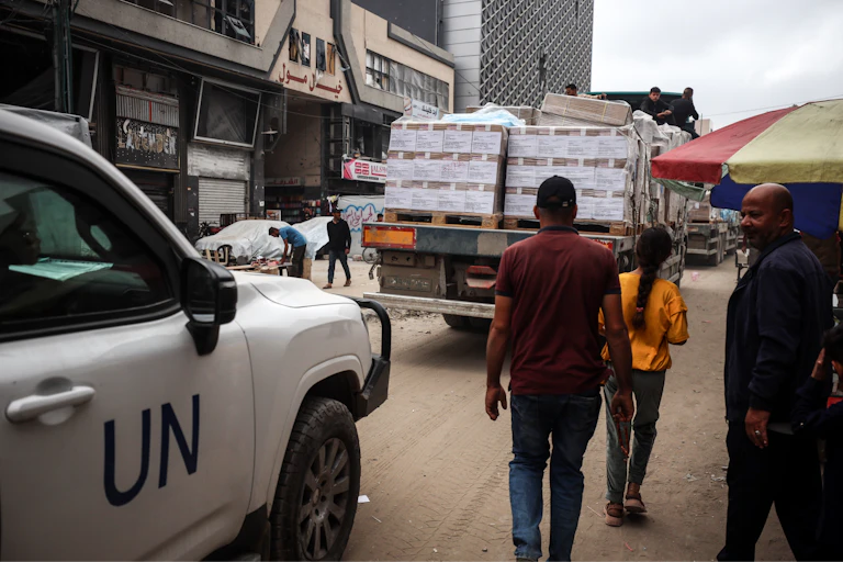 People stand behind a truck with aid containers. A vehicle with UN on the side is behind them.