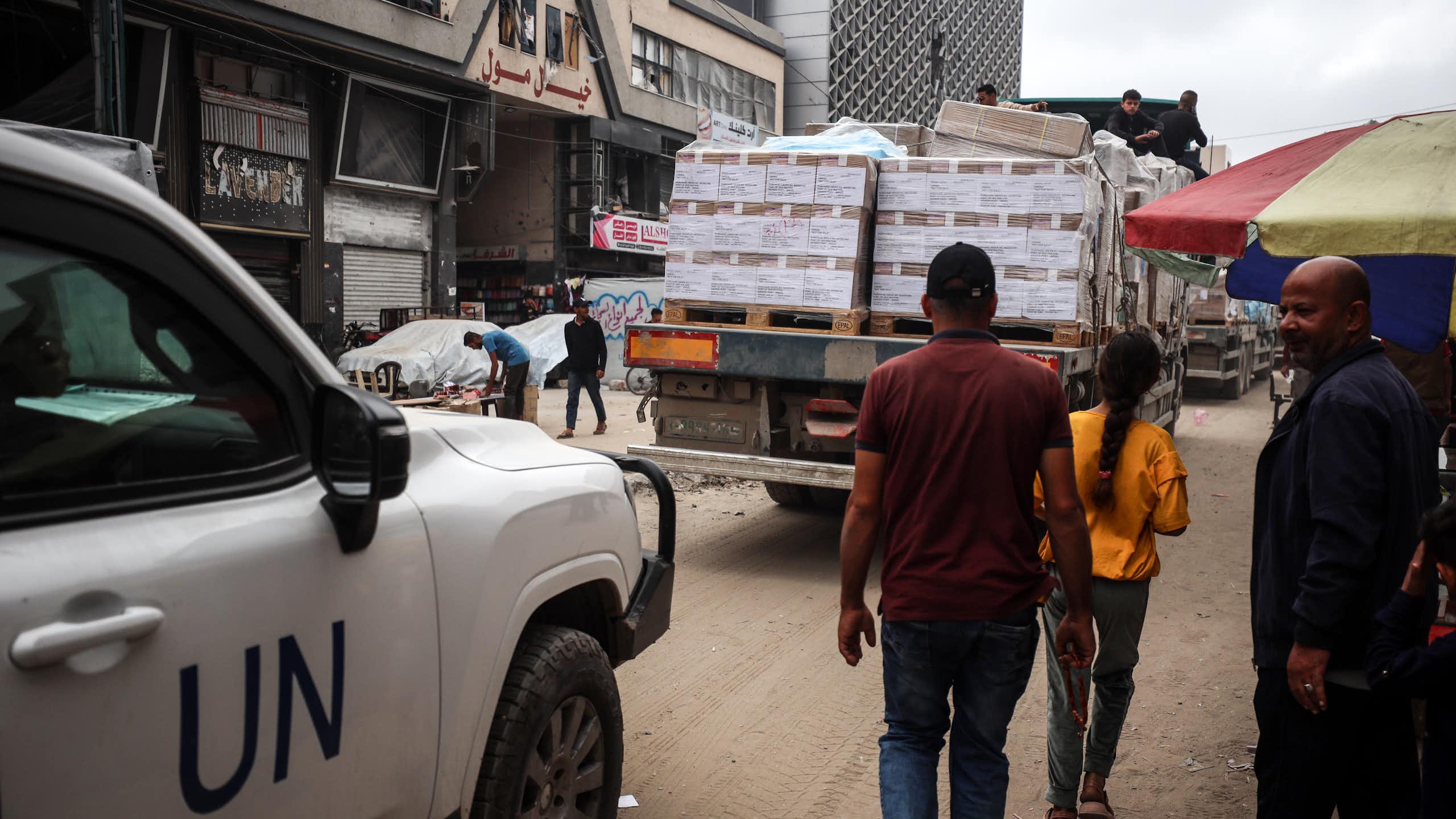 People stand behind a truck with aid containers. A vehicle with UN on the side is behind them.