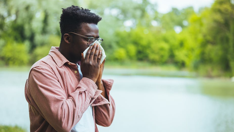 A man with hay fever blows his nose into a tissue while standing in an outdoor park.