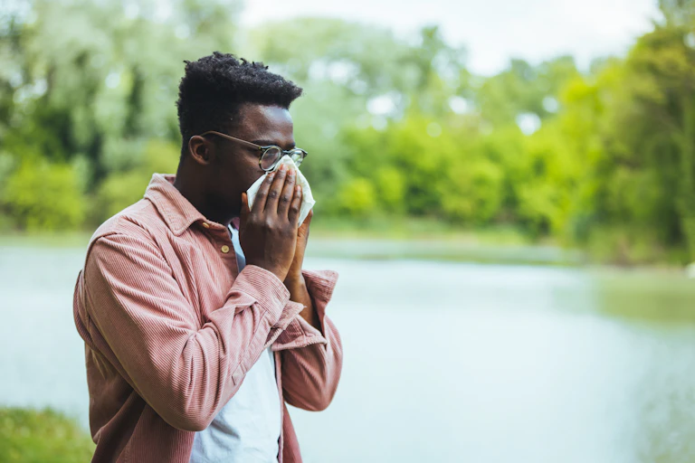 A man with hay fever blows his nose into a tissue while standing in an outdoor park.