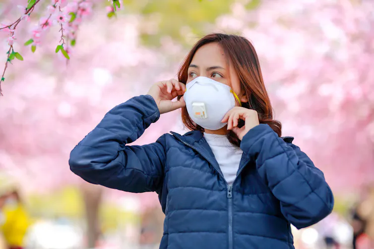 A woman wearing a face mask stands by a row of blossom trees outdoors.