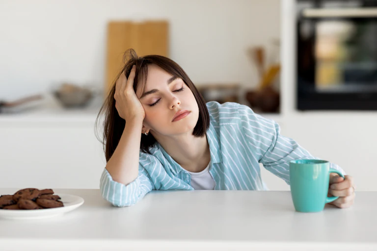 A young woman falling asleep in a kitchen, a cup of coffee in her hand.