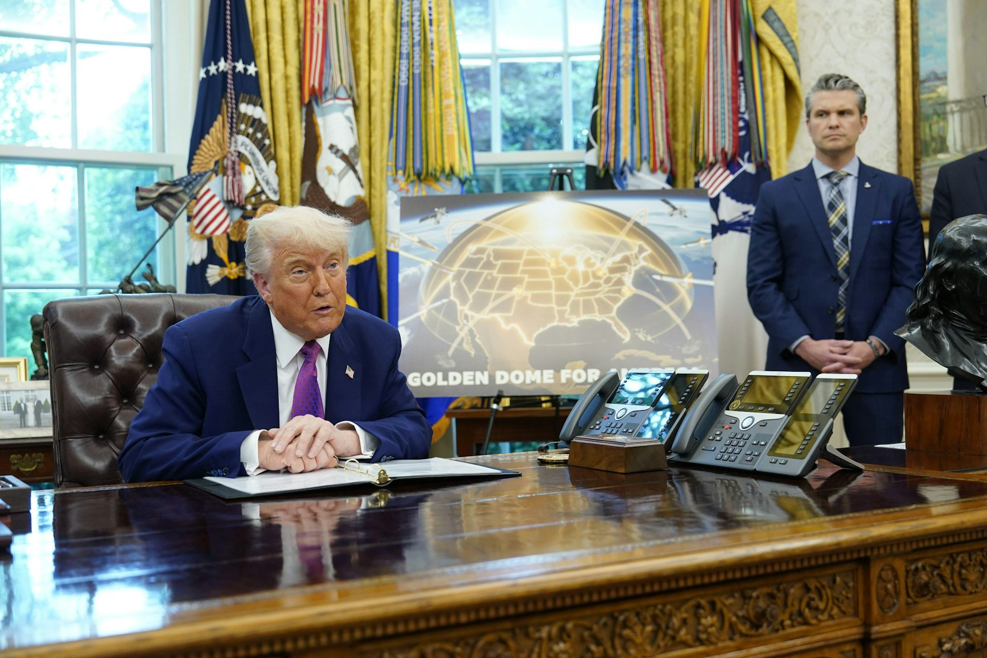 Donald Trump at his desk in the Oval Office in front of a diagram showing a missile defence system with defense secretary Pete Hegseth standing in the background.