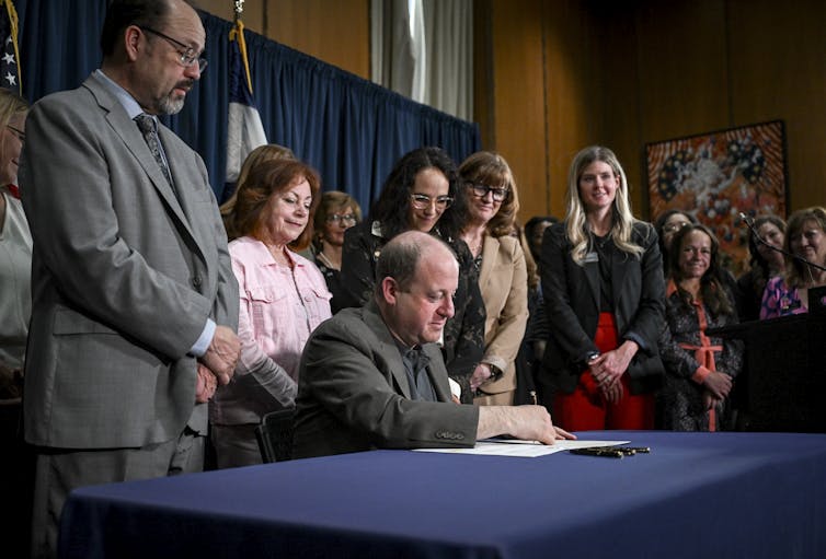 Colorado state lawmakers stand around the Colorado governor who is seated at a table and signing papers.