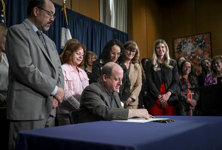 Colorado state lawmakers stand around the Colorado governor who is seated at a table and signing papers.