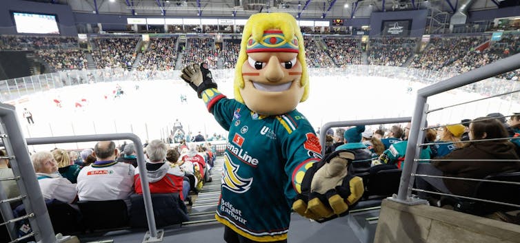 A mascot of a blonde man in a headband and jersey stands in a hockey arena among spectators.