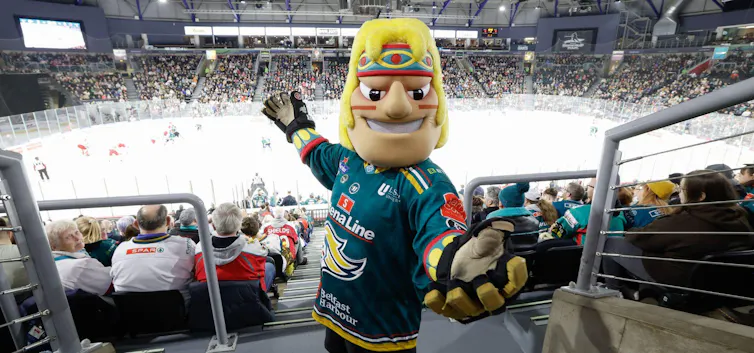 A mascot of a blonde man in a headband and jersey stands in a hockey arena among spectators.