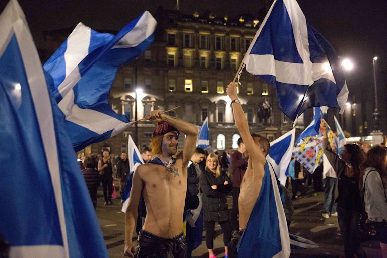 Bare-chested men wave blue and white Scottish flags in a crowded city square.
