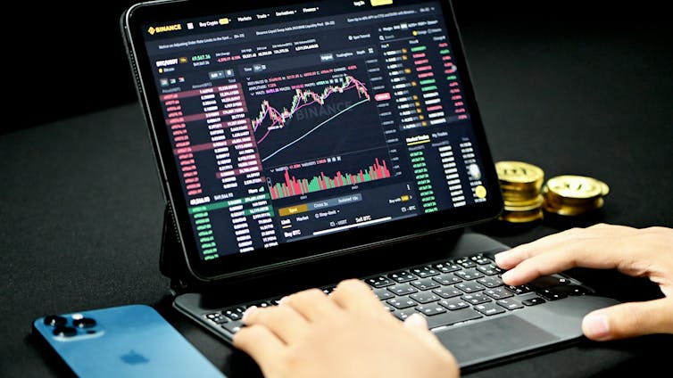 An iPad screen shows a trading platform; hands are on the keyboard. There is a cellphoe to the left and gold coins to the right.