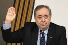 A man with thinning dark hair in a suit and tie with his right hand raised.