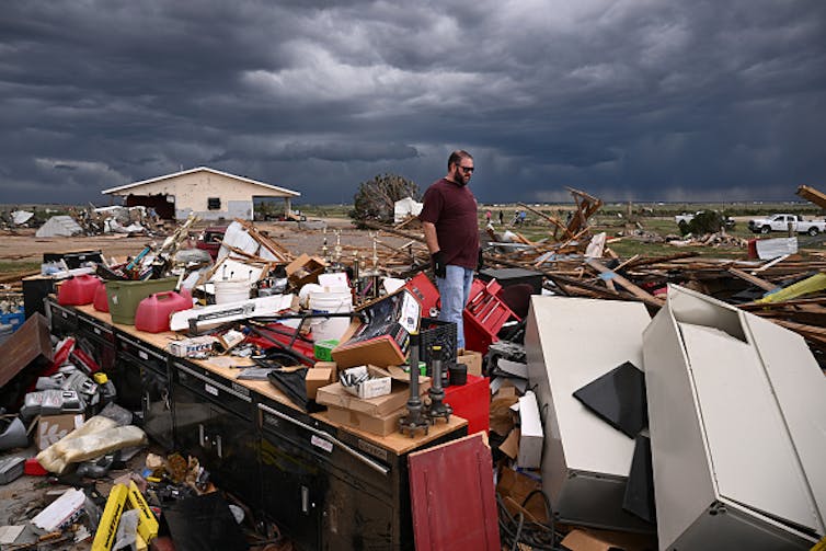 A man stands amid splintered wood. Only a row of cabinets is still standing.