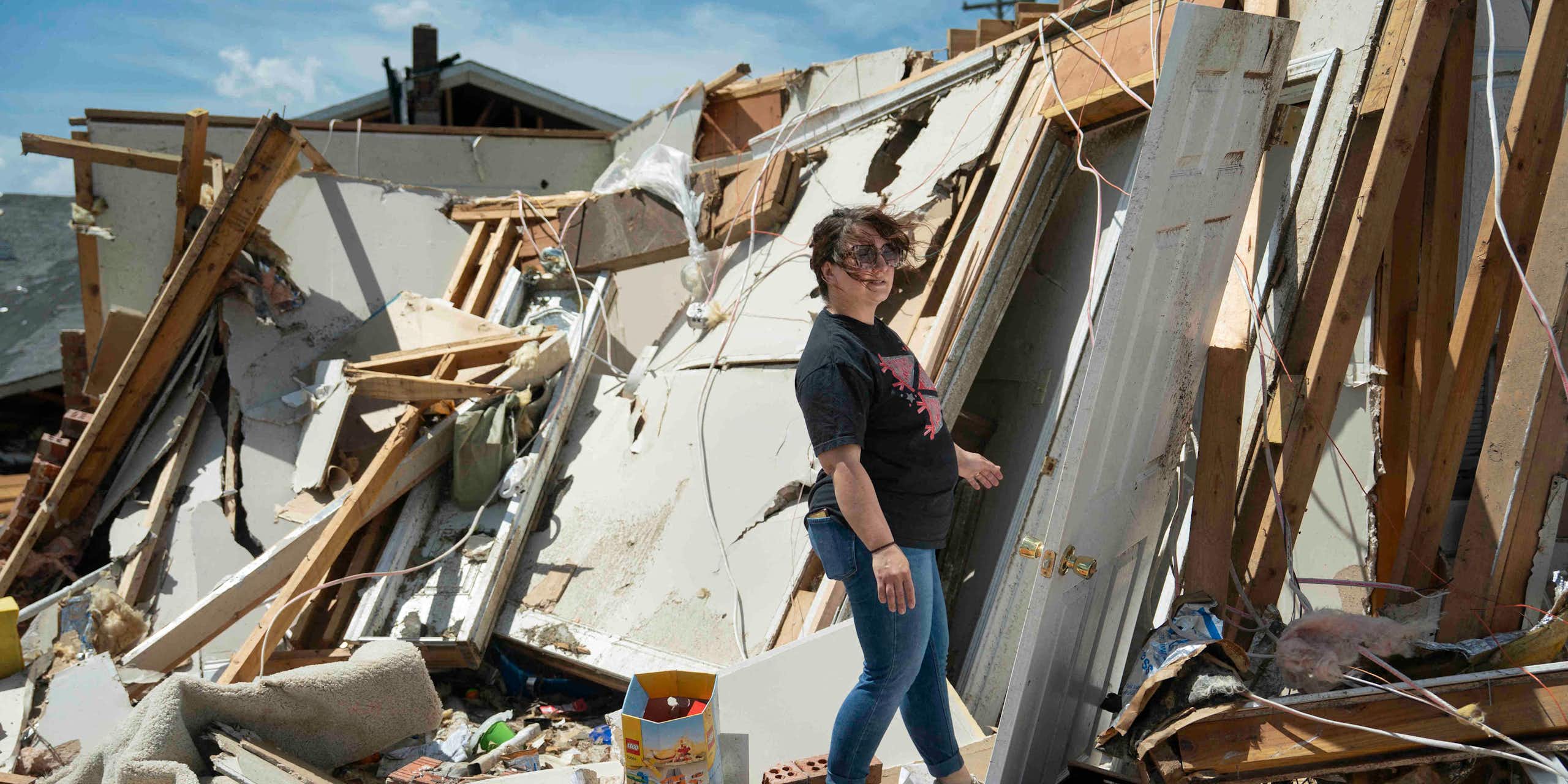 A woman walks through the shredded remains of a home. One wall with a white door is leaning away from her and nothing else is standing.