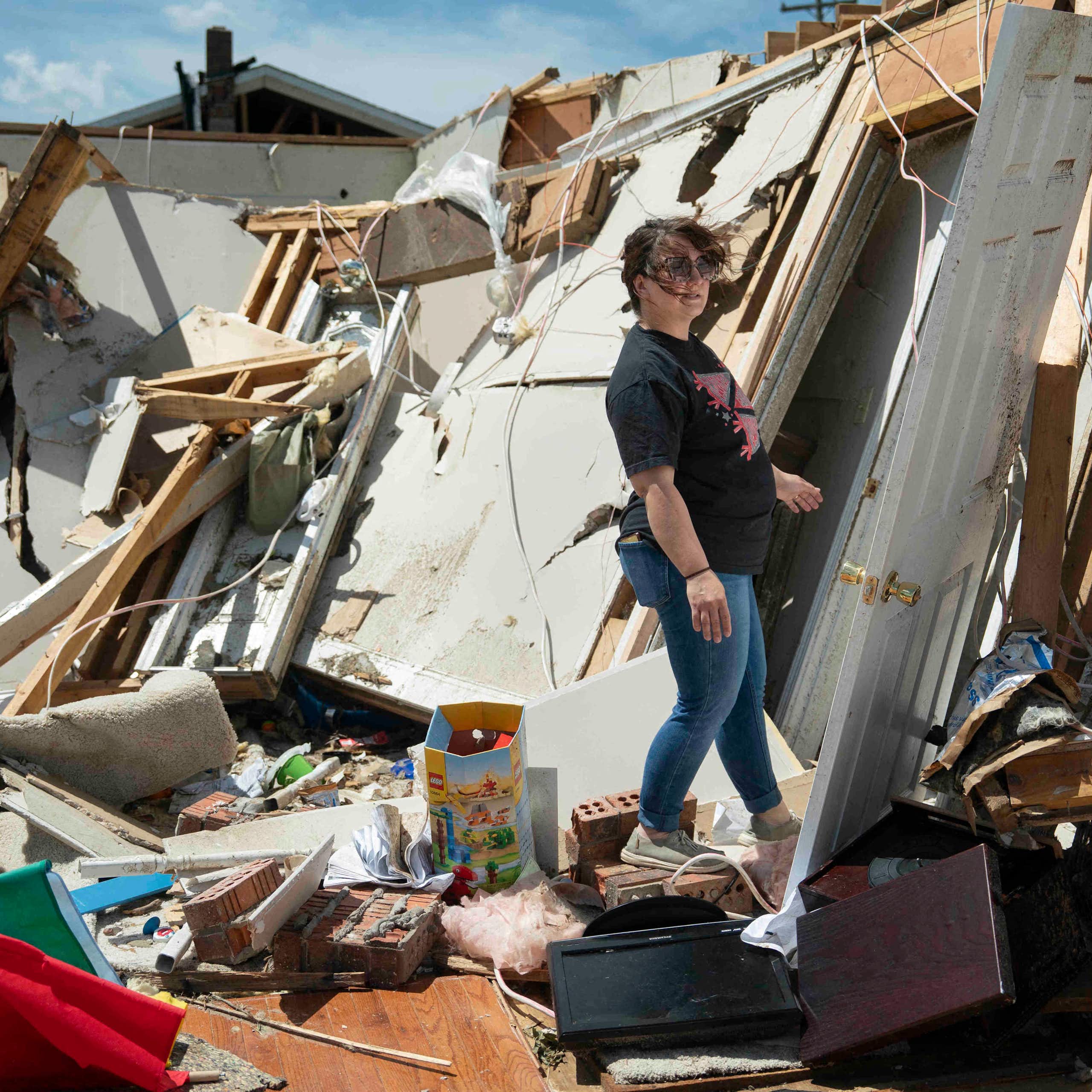 A woman walks through the shredded remains of a home. One wall with a white door is leaning away from her and nothing else is standing.