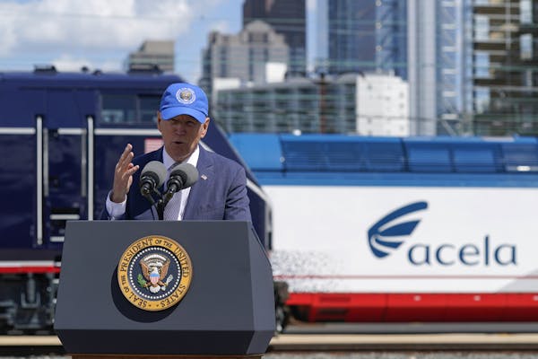 Man in suit and blue baseball cap speaks behind a lectern in front of a train with an urban skyline in the background