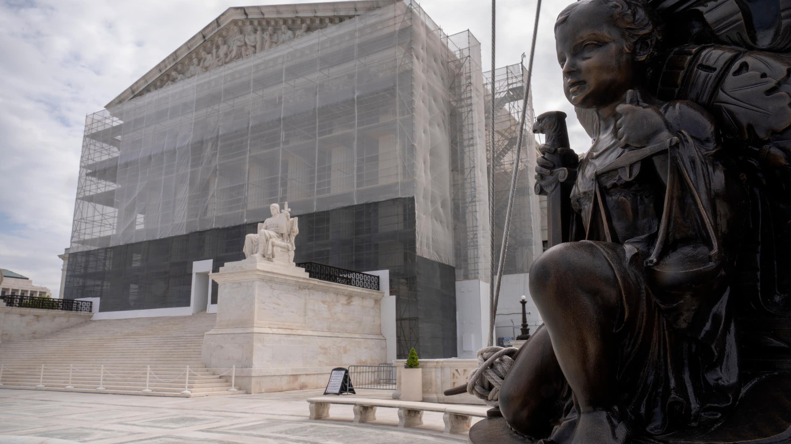 A bronze-colored statue of a child holding scales positioned in front of a large white building with scaffolding around it.