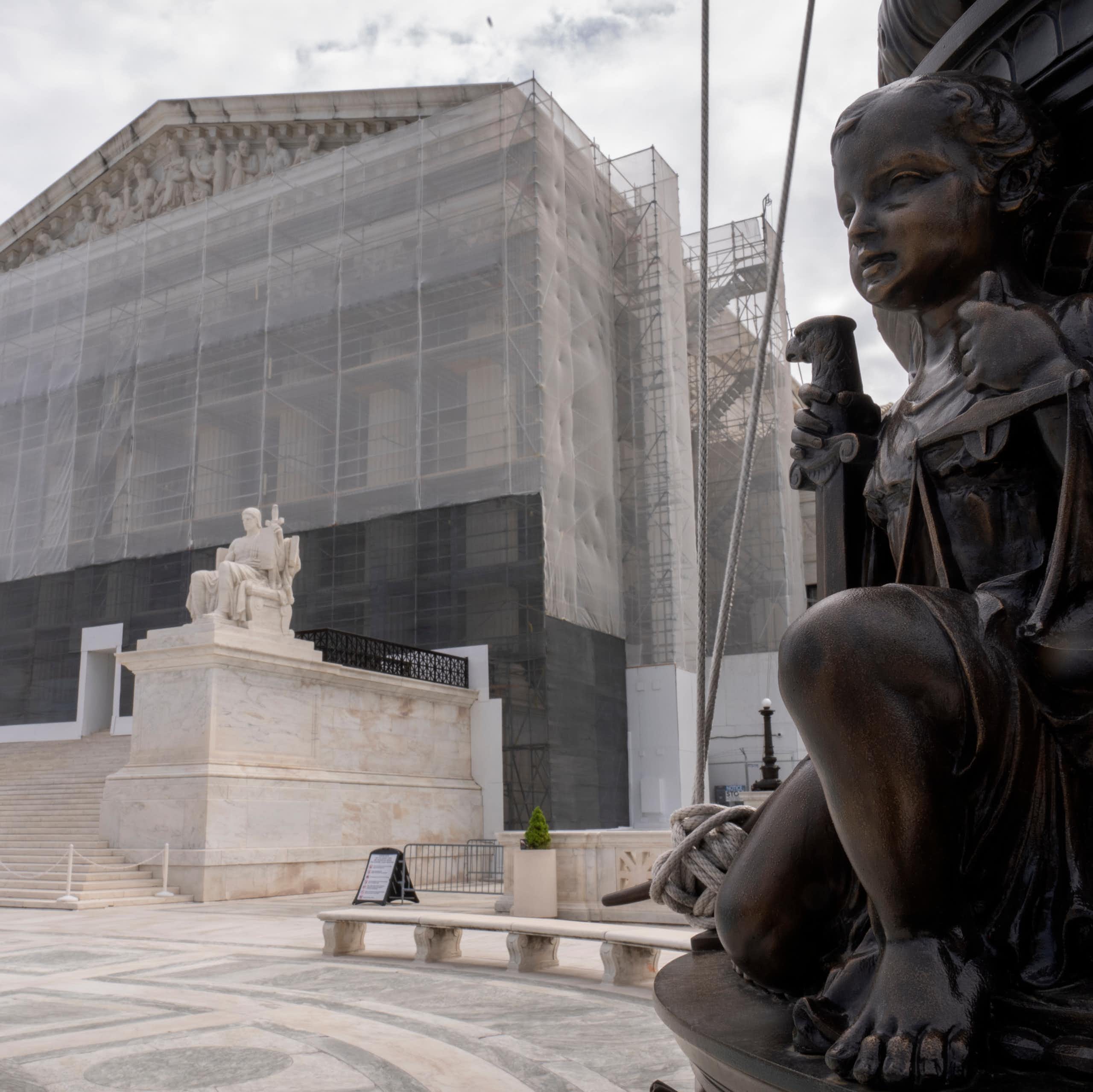 A bronze-colored statue of a child holding scales positioned in front of a large white building with scaffolding around it.