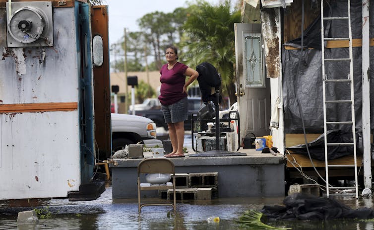 A woman stands on a porch of a home damaged by the storm and surrounded by flooding.