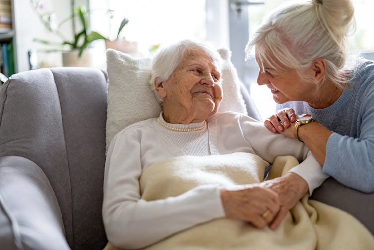 A younger woman interacts with a senior woman in a nursing home. Both look happy.