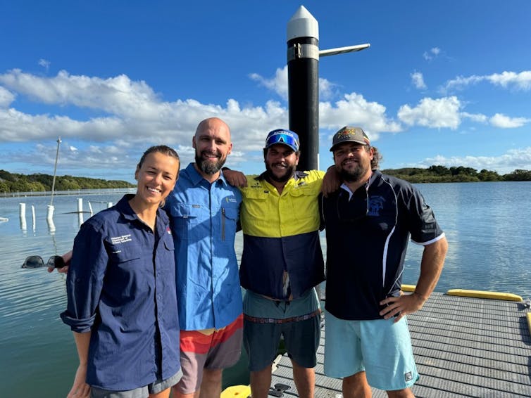 A group of four people standing with their arms around each other on a jetty with an estuary in the background.