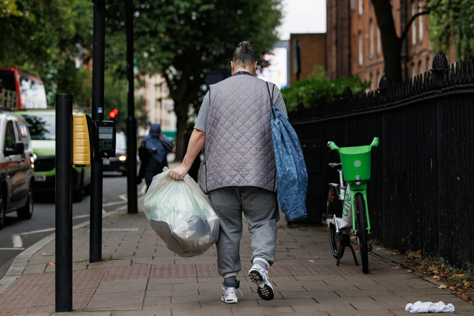 View from behind of a man in gray clothing walking down a street, carrying a plastic bin bag full of belongings