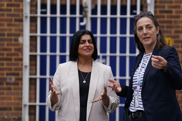 Mahmood standing in front of a barred gate, speaking with prison governor Sarah Bott who is gesturing towards the camera