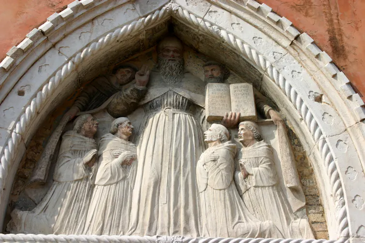 A relief depicts a tall, bearded man holding an open book, flanked on either side by two kneeling men.