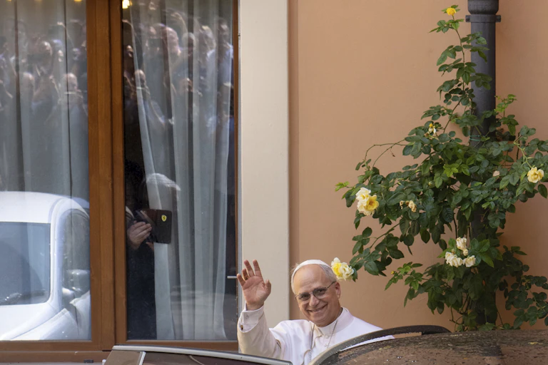 A man dressed in white priestly vestments waves with a smile.