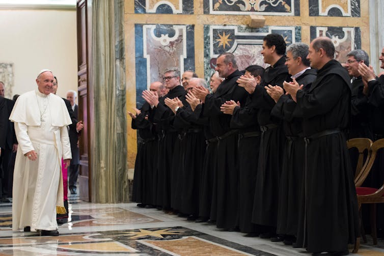 A man in white priestly vestments greets a group of men dressed in black clerical garments, who applaud as he walks in.