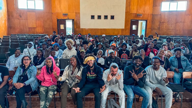 Hip hop artists sitting in a crowd of smiling young people in an auditorium.