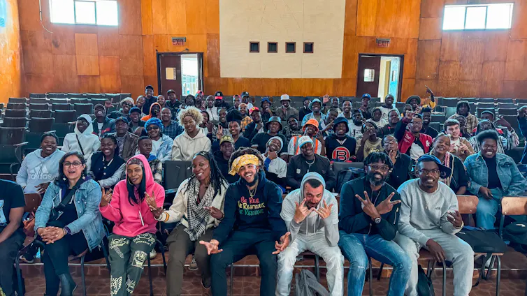 Hip hop artists sitting in a crowd of smiling young people in an auditorium.