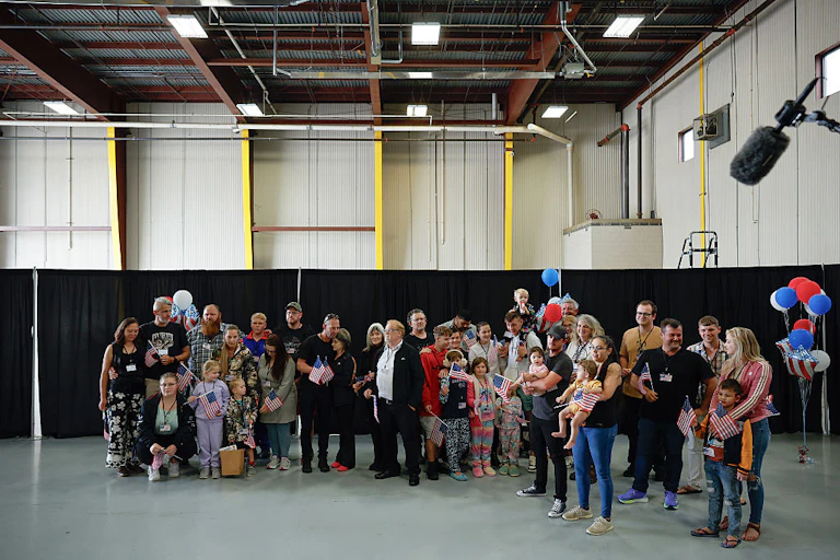 A group of adults and children standing in a large indoor space, with some balloons in the background. Some are holding small US flags.