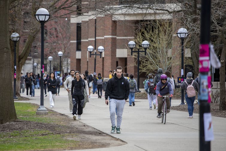 Young people walk and bike along a paved road lined with red brick buildings on one side and trees on the other.
