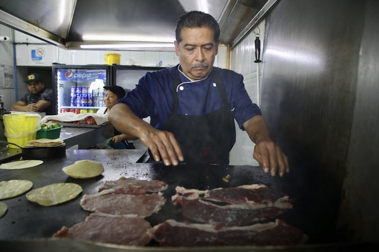 A man in blue uniform and black apron places thin slices of meat on a griddle