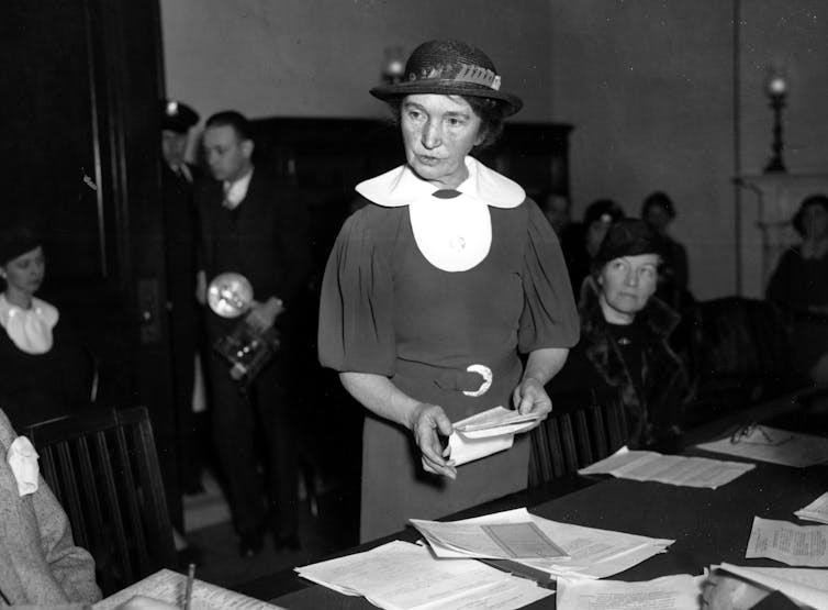 A woman in a hat speaks in front of a table with papers.