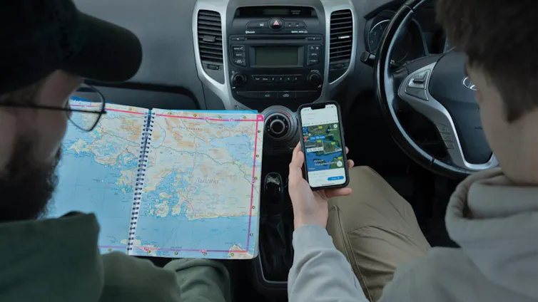 Two people seated in a car, one holding a map open, and the other with a map app on their phone
