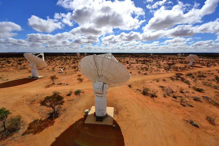 An overhead view of large white radio dishes under a bright blue sky littered with clouds and a red earth underneath.