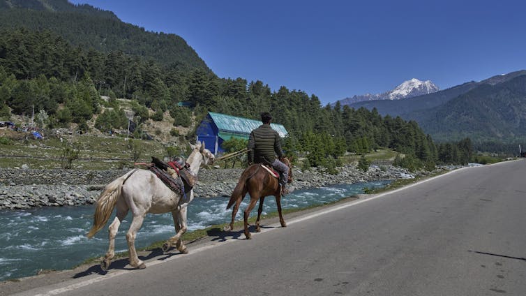 Mountains in the background, a river in the foreground - next to river, a man rides a brown horse leading a pony by a rope along the river