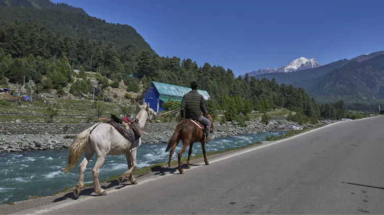 Mountains in the background, a river in the foreground - next to river, a man rides a brown horse leading a pony by a rope along the river