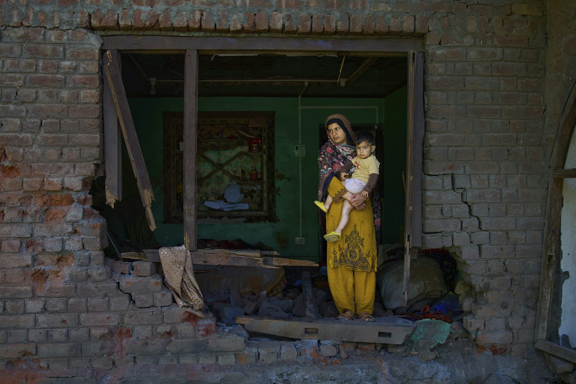 a woman in a yellow sari stands wth her baby looking through a large whole in her brick house 