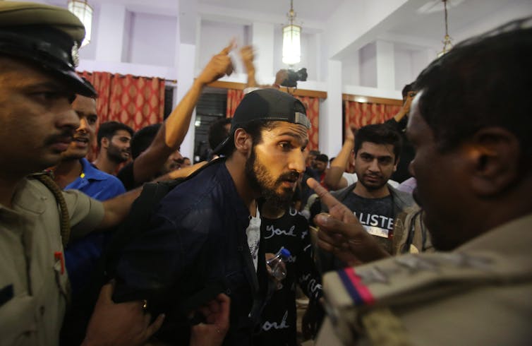 A thin man dressed in black with trim beard is surrounded by men in khaki police uniforms with caps.