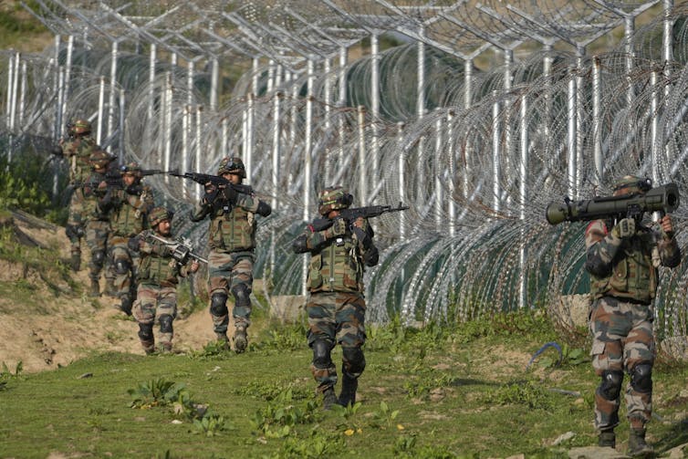 Solders in green and helmets holding and pointing weapons walk along a high barbed wire fence