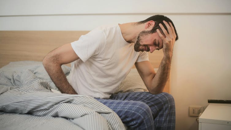 A man sitting on the edge of a bed holding his forehead.