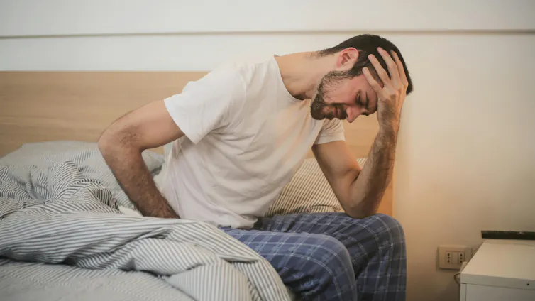 A man sitting on the edge of a bed holding his forehead.