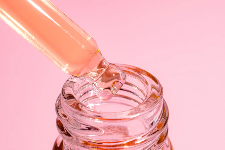 Pipette drips into a glass bottle on a pink background.