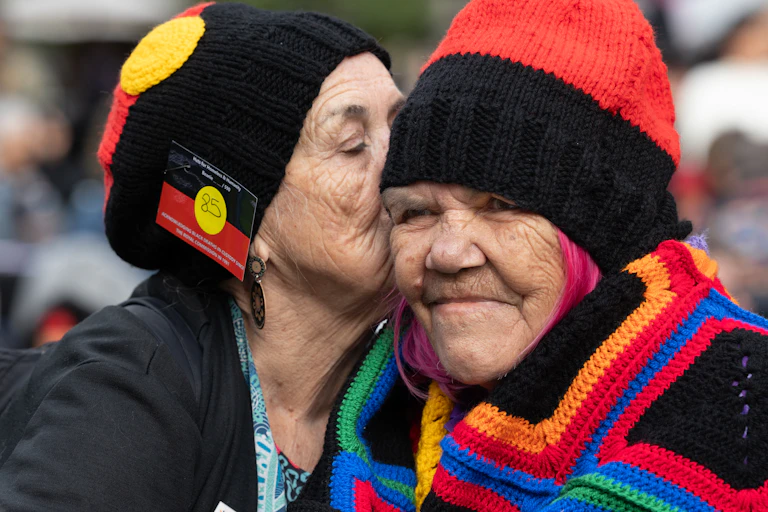 Two older First Nations women wearing black, red and yellow beanies.