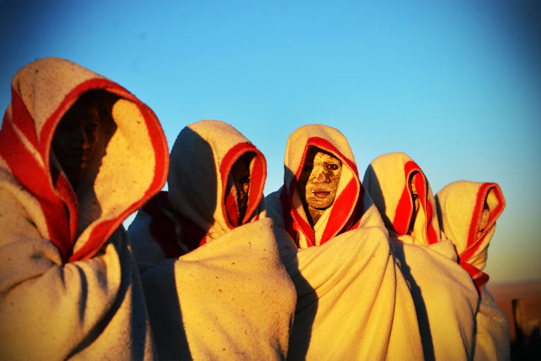 A group of five stand with blankets wrapped around themselves, faces smeared with mud are barely visible.