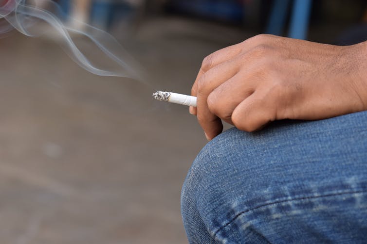 A man holds a lit cigarette in his hand, which rests on his knee.