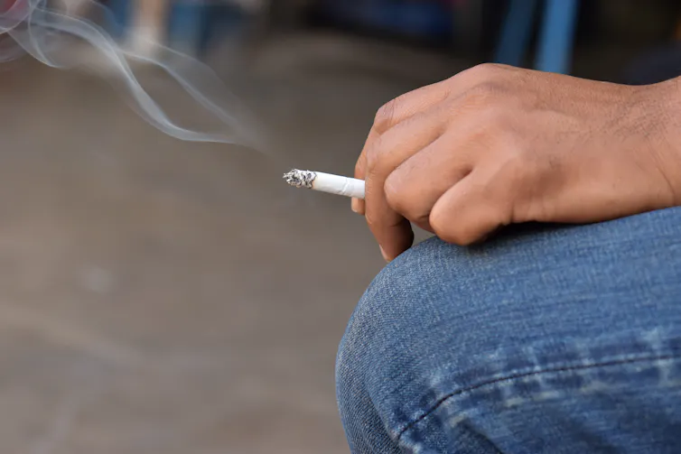 A man holds a lit cigarette in his hand, which rests on his knee.